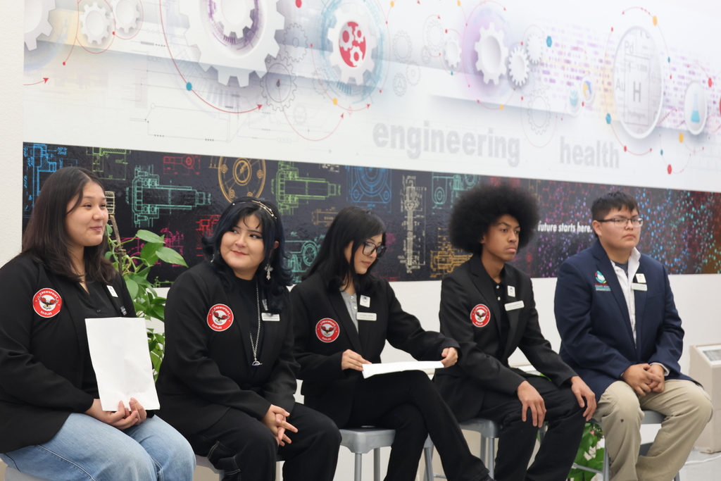 Five people in black jackets sit on chairs, holding papers. One wears a suit. Behind them, a banner reads "engineering health."