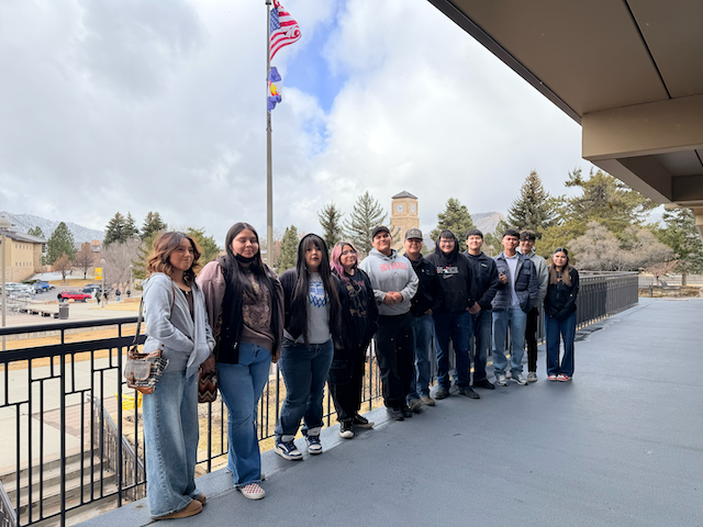 A group of people stands on a balcony with railings. They are wearing jackets and jeans. Behind them, a flagpole and trees are visible.