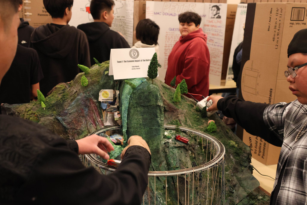 Several students view a model mountain with a road, tunnel, and tunnel exit. Some wear glasses.