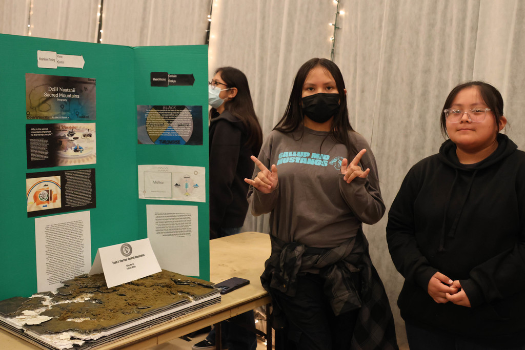 Three people stand near a green poster board displaying a project. One person poses with fingers crossed.