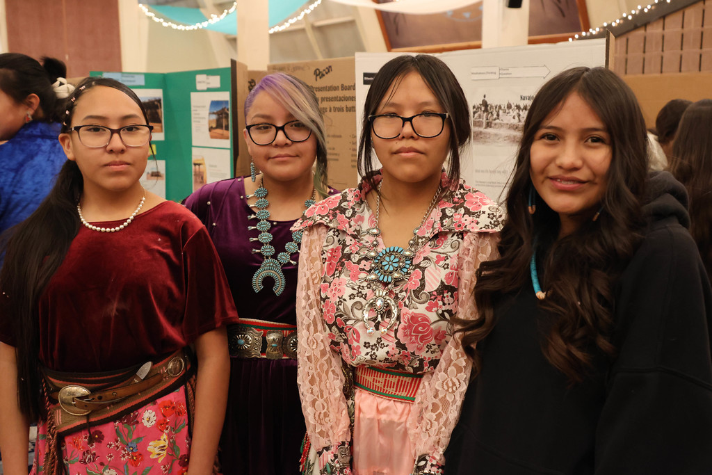 Four women in traditional clothing pose for a photo in a room with posters on the wall.
