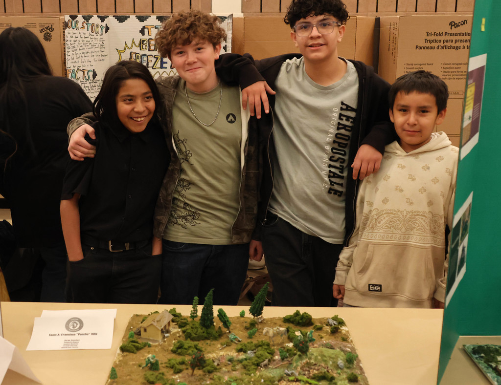 Four kids pose next to a miniature landscape model at a school event. Behind them, storage boxes and other displays are visible.