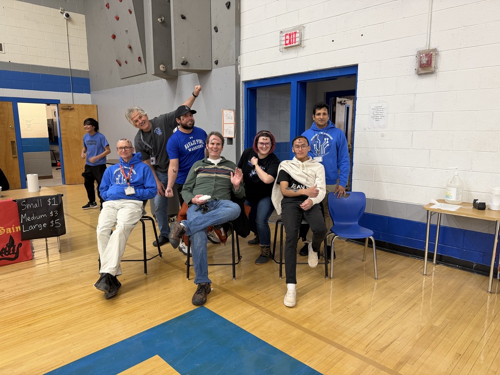 Group of people seated in a gym, some smiling, with tables and chairs in the background.