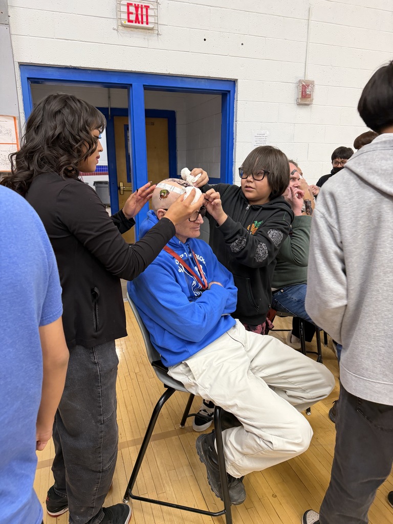 People in a room. A person sits on a chair, and another person adjusts a mask on their head.