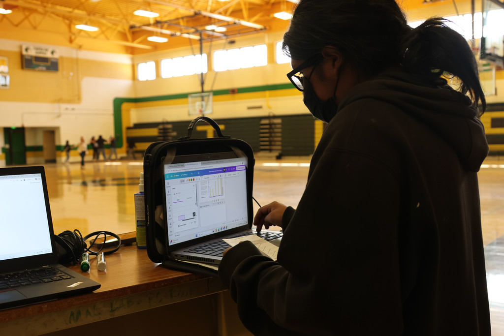 Person in a gym with two laptops, one open, wearing a mask and glasses, standing behind a table.