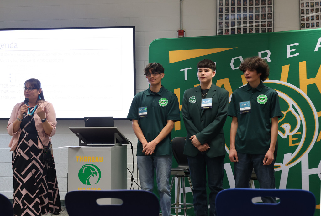 Four individuals in green shirts stand before a screen. A woman in a black and white dress stands to the left.