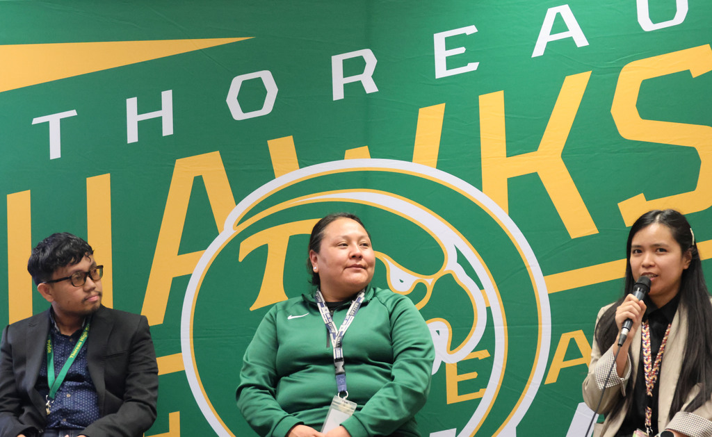Three individuals seated in front of a green backdrop. One holds a microphone, while the others sit behind her.