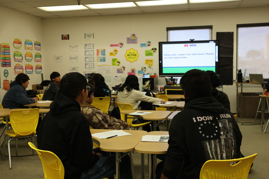 Students seated in a classroom with yellow chairs and tables. A projector screen displays information.
