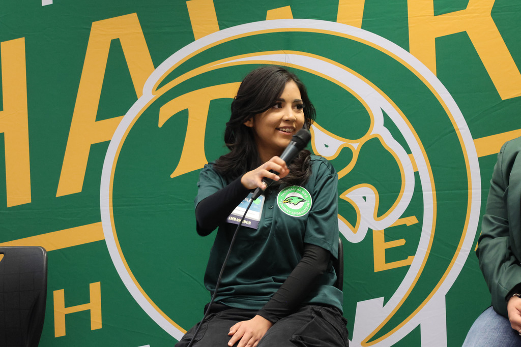 Woman seated, holding a microphone, speaking in front of a green and yellow backdrop with text.