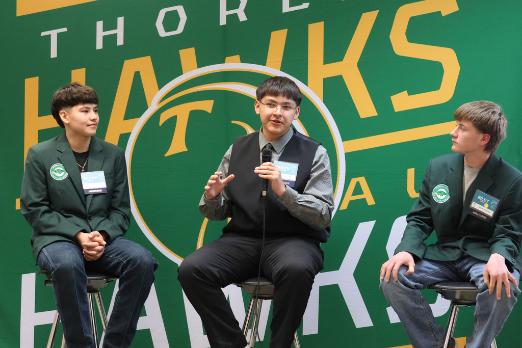 Three young men in green jackets sit on stools in front of a green sign.