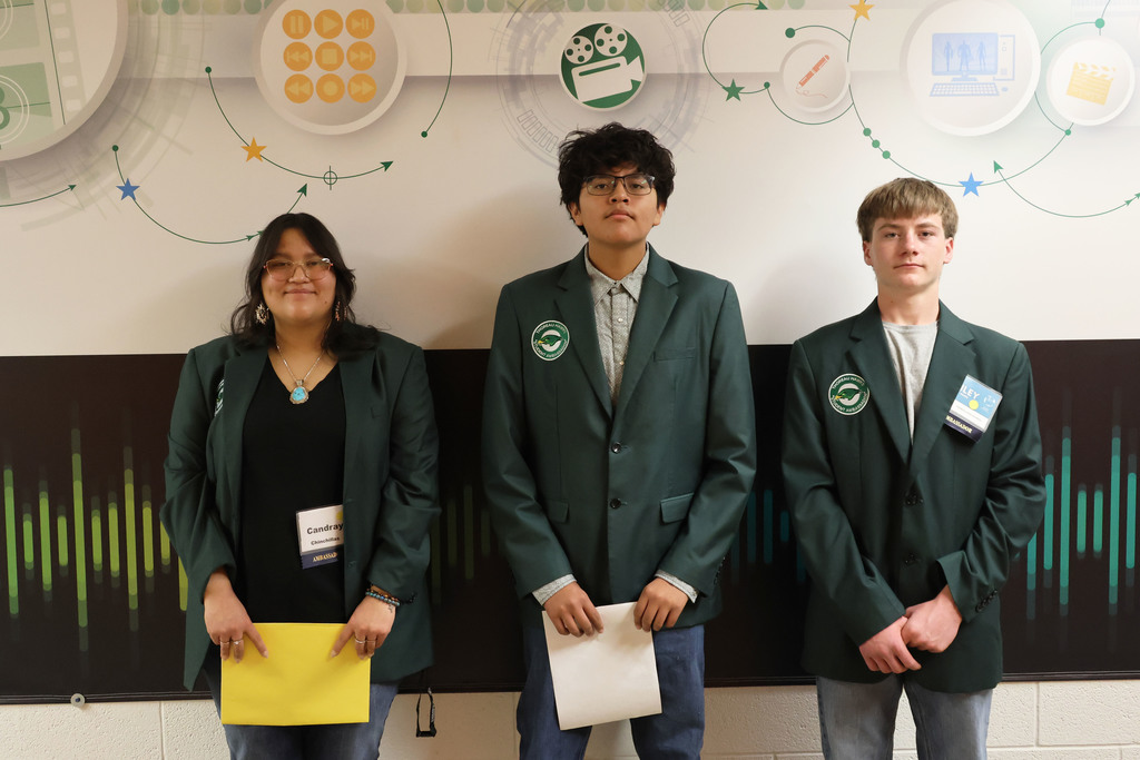 Three individuals in green coats stand together in front of a colorful wall with icons.