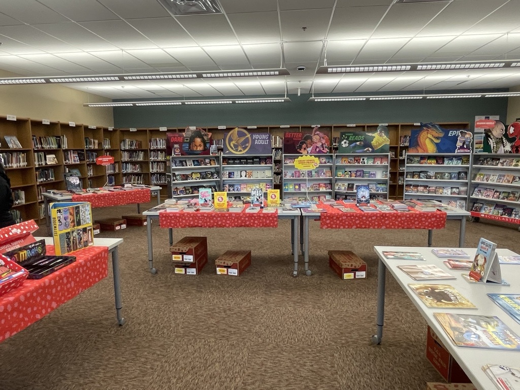 Library with rows of bookshelves and long tables covered in red fabric, displaying books. A person stands at the left.
