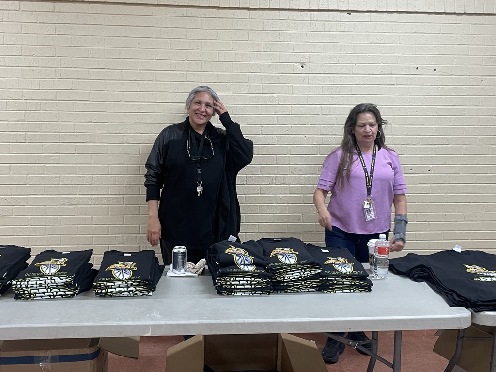 Two women stand at a table with several black T-shirts, one woman holds her head.