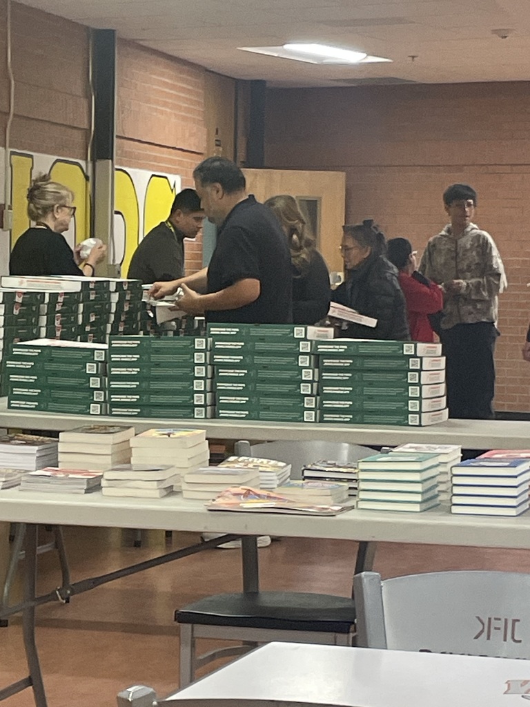 a table with stacks of books, a table with stacks of pizza boxes, people in a line behind tables
