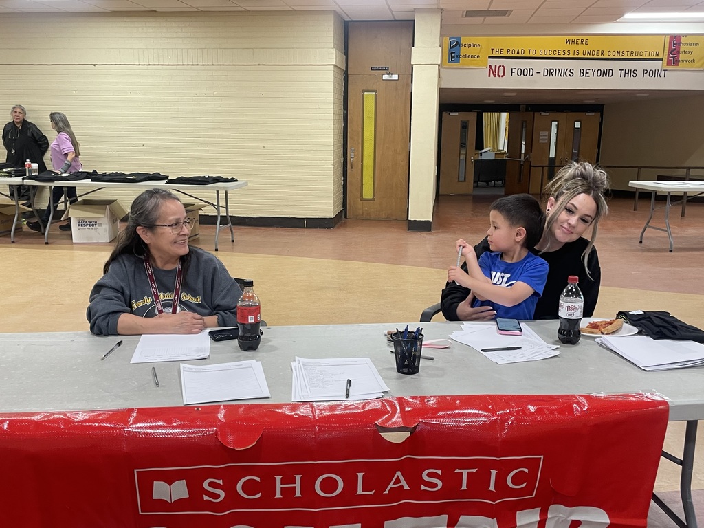 Three people sit at a table with Scholastic banners. A woman holds a child, while another woman sits with glasses.