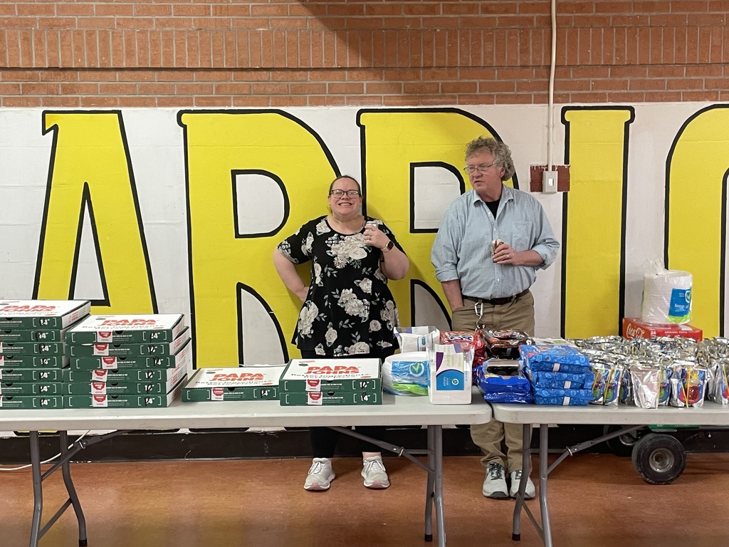 Two people stand behind a table filled with boxes and products in front of a yellow sign.