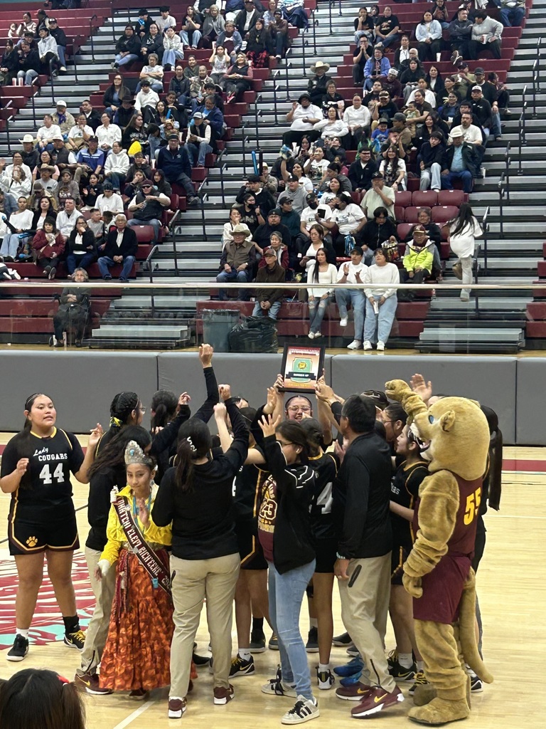 People celebrate in a gymnasium. Some hold a trophy. A mascot stands among them. Spectators sit in the stands.