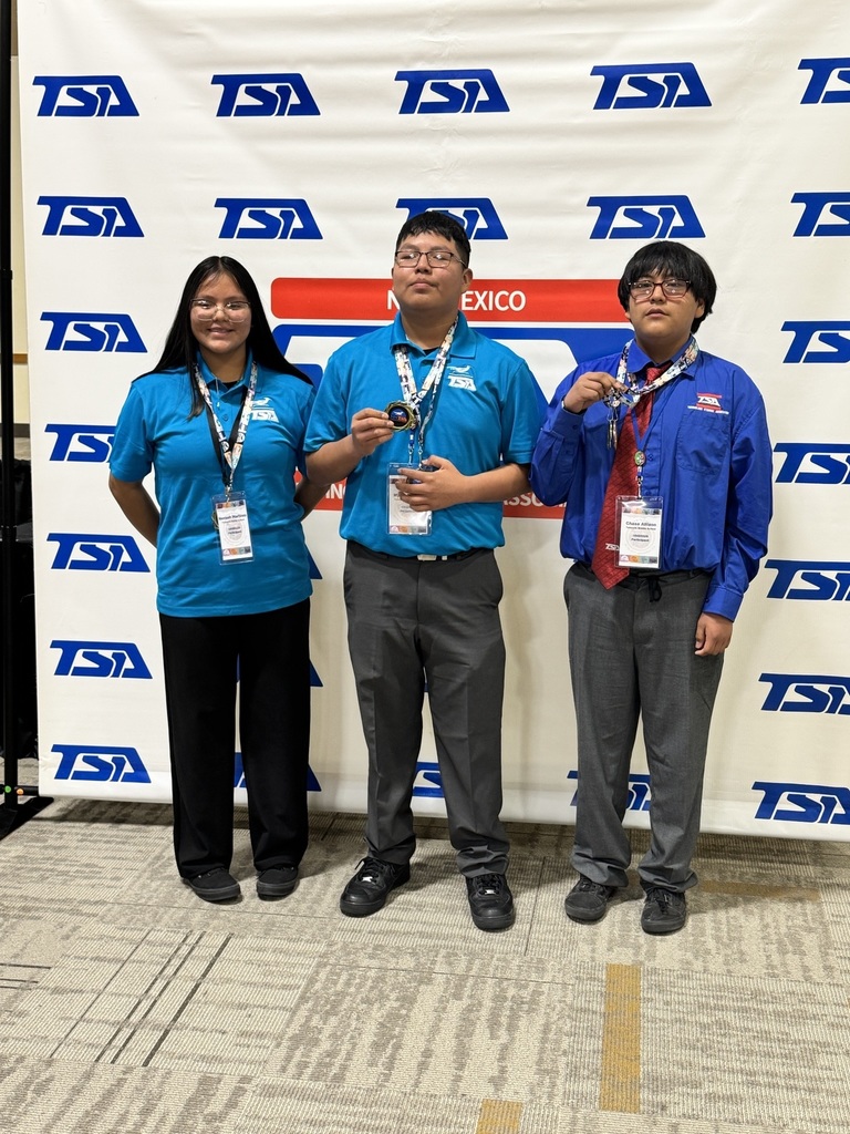 Three people stand together in front of a banner with large blue letters. They wear light blue shirts and hold medals.
