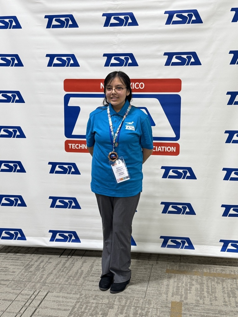Person in blue shirt with TSA logo stands in front of a white banner. Floor has gray tiles.