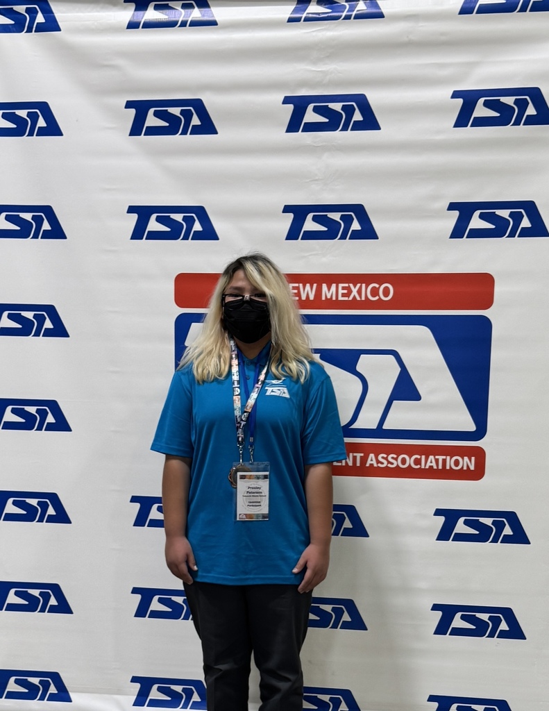 A person with blonde hair stands in front of a backdrop featuring blue, red, and white text.