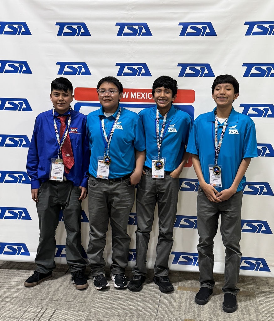 Four boys in blue shirts and gray pants pose together. They stand before a banner with large blue letters.