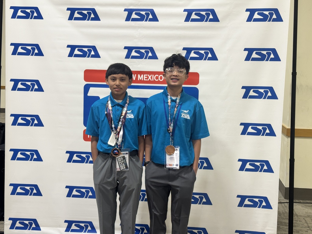 Two young boys in matching blue shirts and gray pants stand before a white banner with blue text.