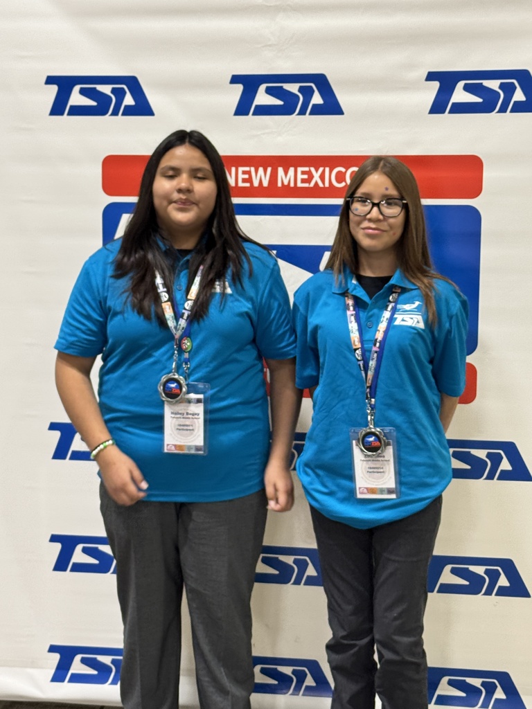 Two women in matching blue shirts stand in front of a white backdrop with "TSA" and "NEW MEXICO" on it.