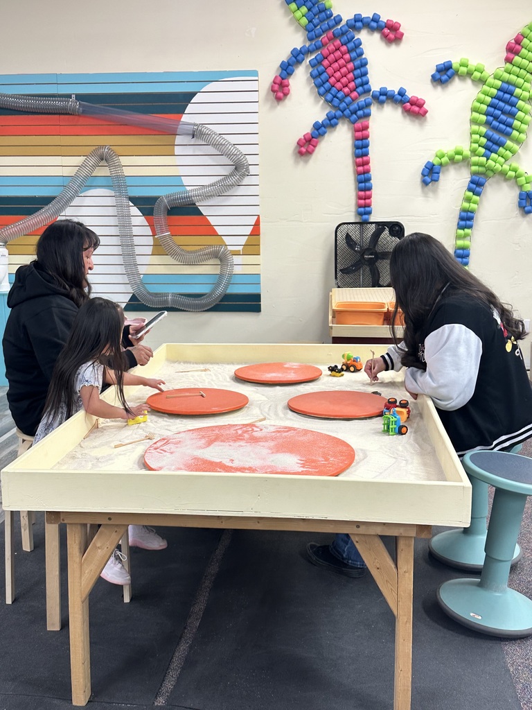 Two adults and a child engage in an activity at a table with orange objects, surrounded by colorful wall art.