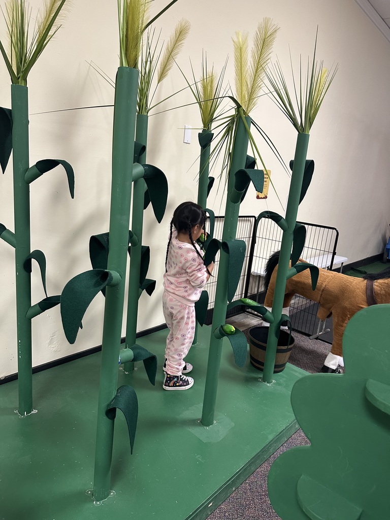 A child wearing pink pajamas plays in a room with corn stalks and a dog.