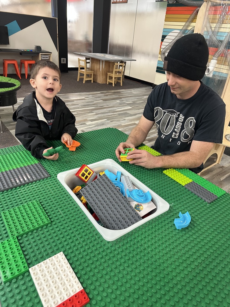 A man and a child are building with colorful Legos on a green table in a playroom.