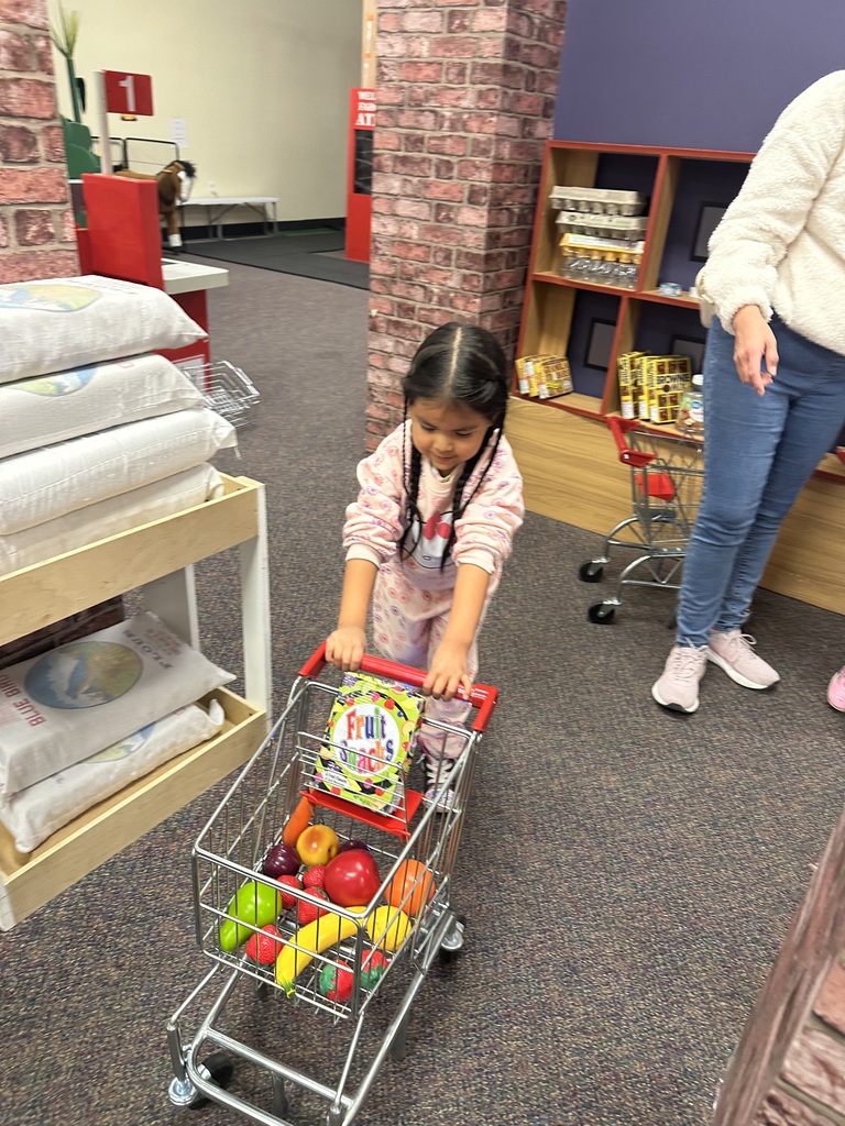 A young girl pushing a toy cart filled with plastic fruits in a play area.