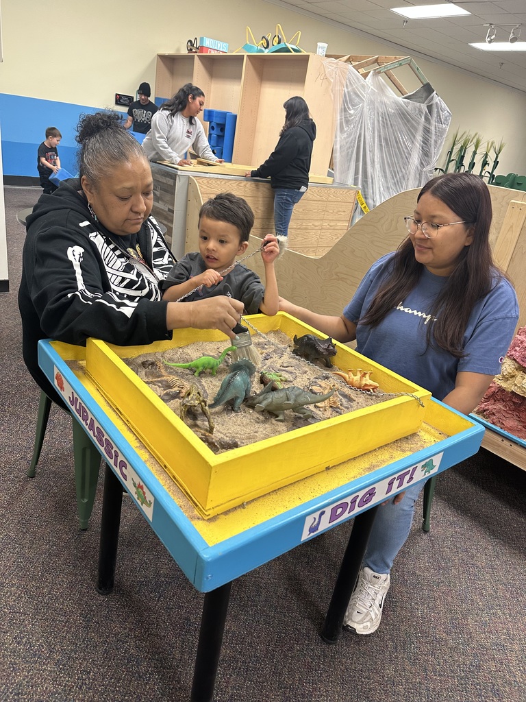 Two adults and a child examine dinosaur toys in a sand tray. Background features a play area with shelves and a partially covered object.