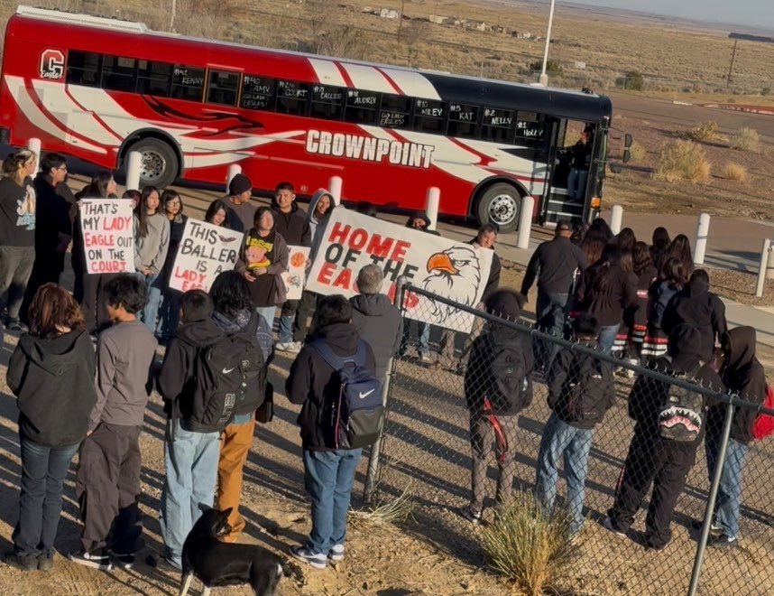 Group of people with backpacks standing by a fence. A red bus is parked in the background.