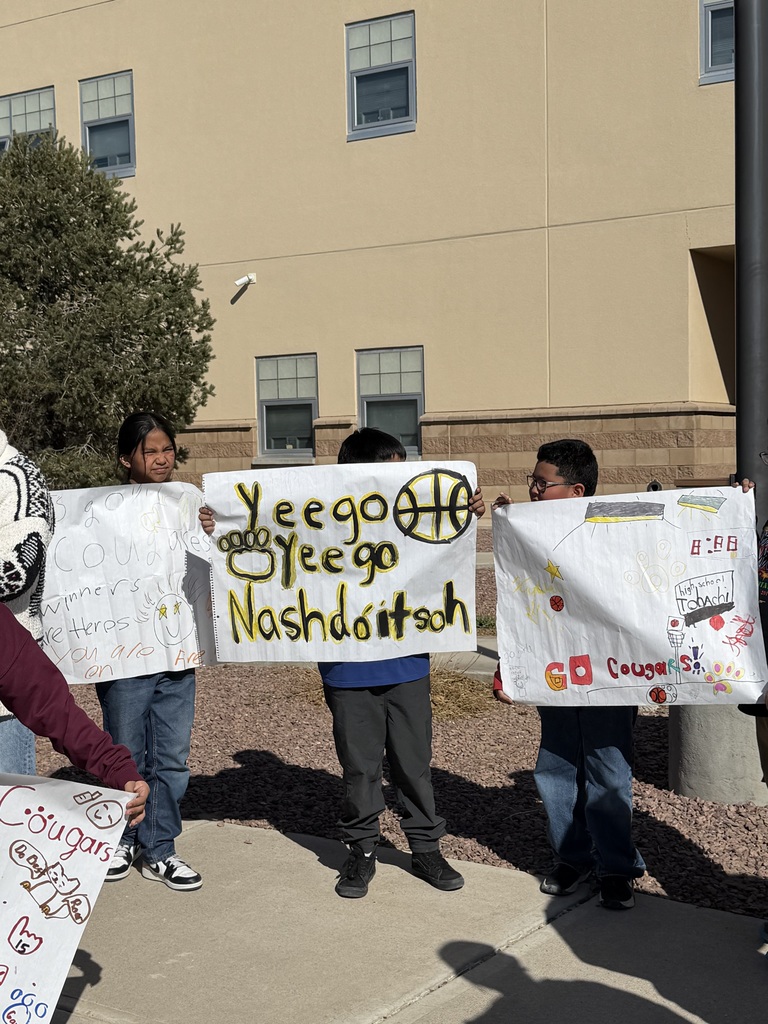 Children holding signs stand on a sidewalk in front of a building with a tree.