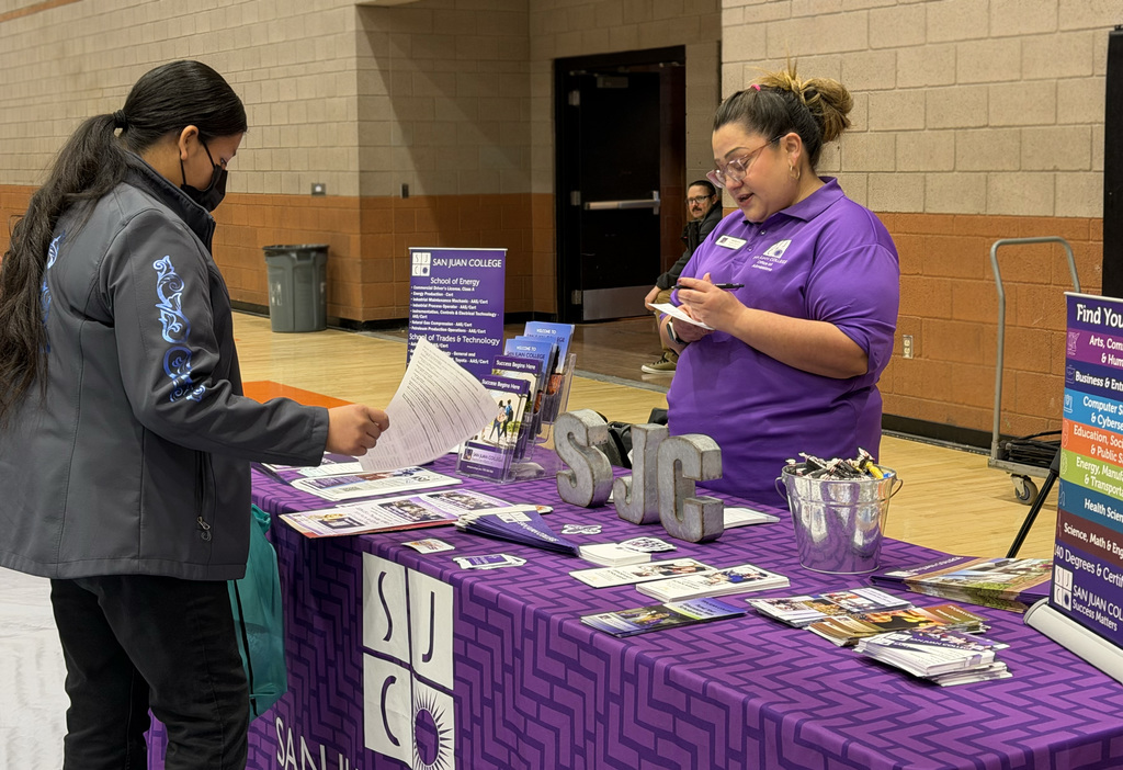 Two women at a table with a purple tablecloth. One holds a paper, the other a pen. A sign reads "SJC" on the table.