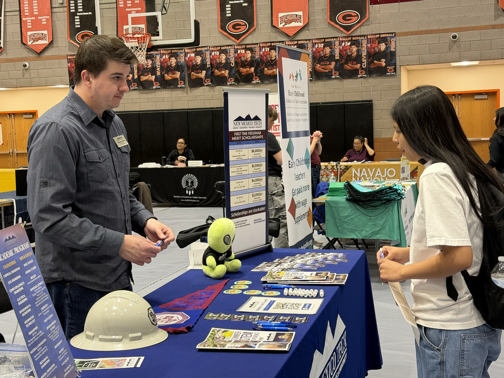 A man and woman converse at a booth with a blue table displaying various materials, helmets, and a stuffed toy.