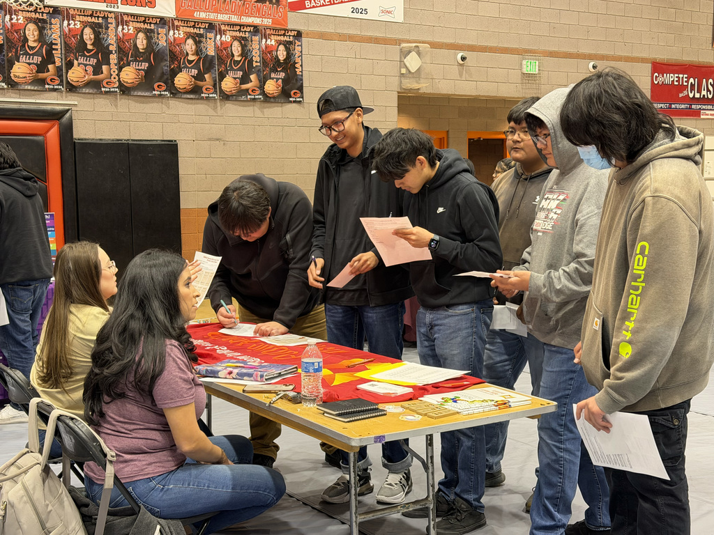 Group of students at a table with documents. Some are seated, others are standing. One student holds a paper.