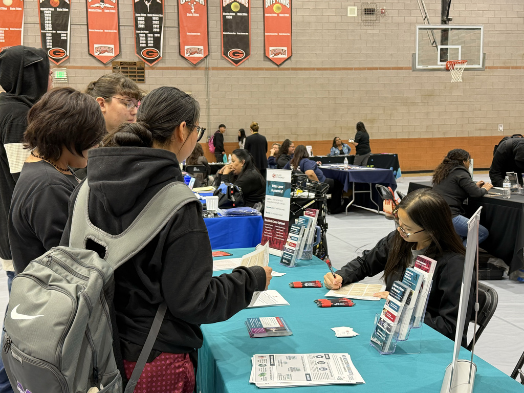 People in a gym are gathered around tables. A woman is seated, writing. A man with a backpack stands and converses with a woman in front of him.