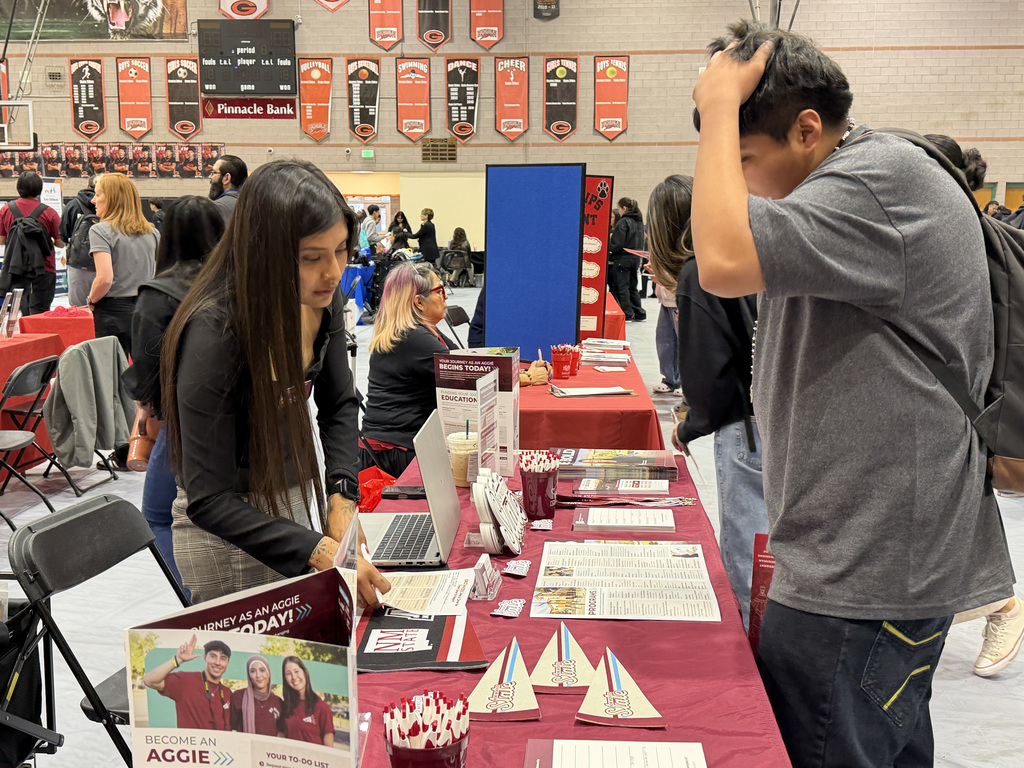 People review information at a college fair. A woman with long hair views a laptop. A man nearby scratches his head.