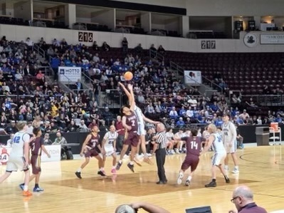 Basketball players in maroon and white uniforms play in an arena with an audience watching.
