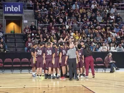 A basketball team huddles on court, facing spectators. Numbers 20, 30, 31, and 2 on jerseys. A referee stands nearby. Audience in stands.