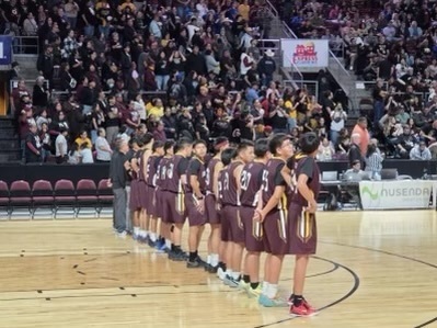 A basketball team stands in line, facing the audience, with a crowd seated in the background.