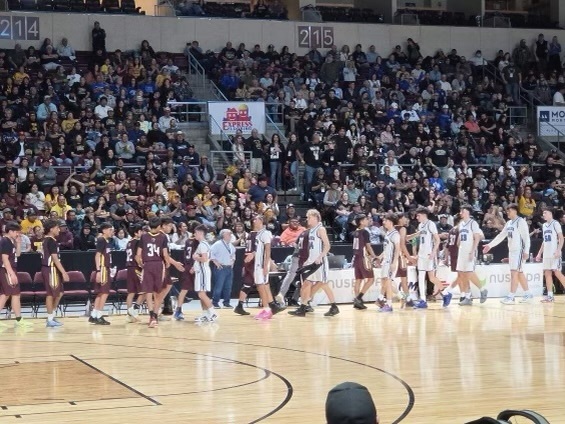 Basketball players in maroon uniforms walk across a court, surrounded by spectators seated in stands.
