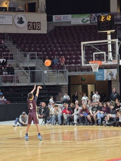 Basketball player in maroon uniform shooting basketball. A crowd of people sits on the bleachers. Digital clock.
