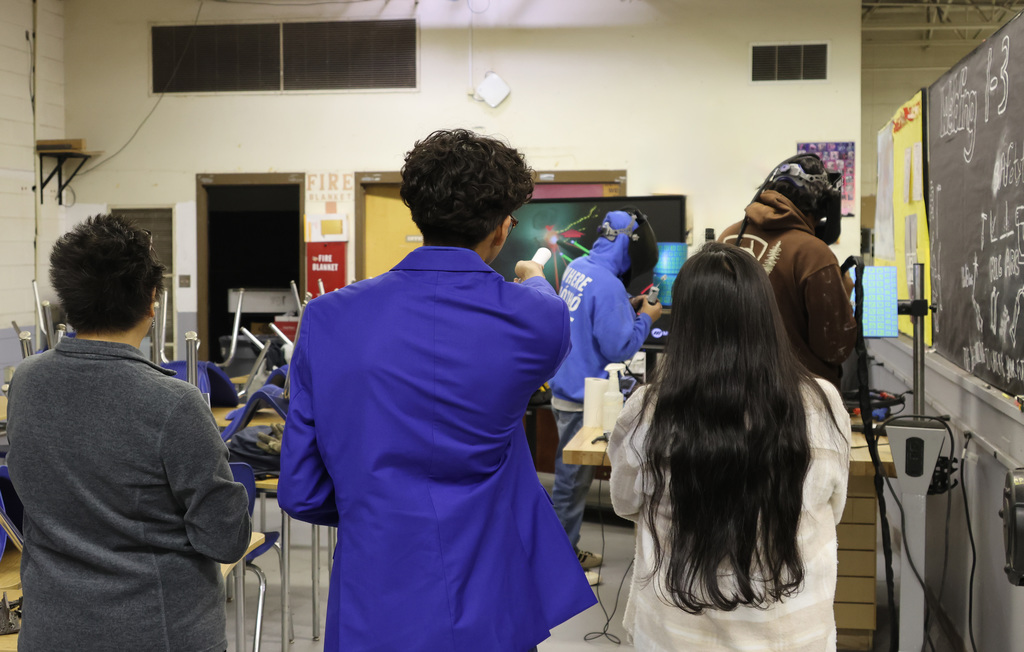 Five people are standing in a room, some wearing lab coats. One person in a blue suit is speaking.