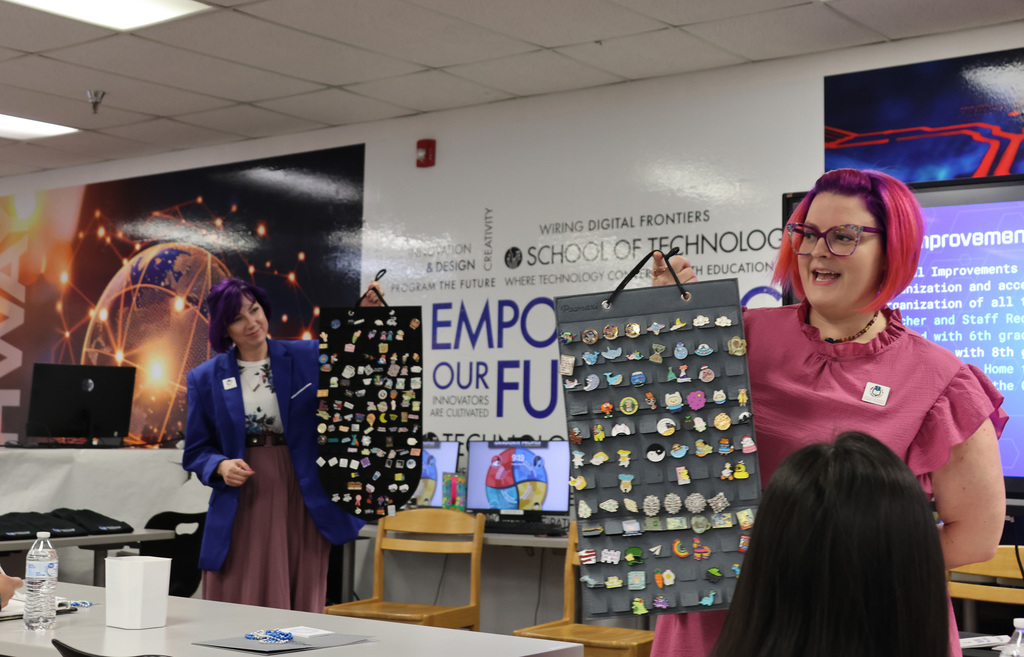 Two women with pink hair hold a board with pins in front of a classroom.