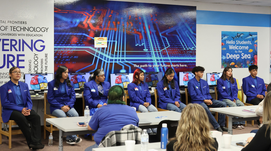 Students sit at tables in a room with a blue circuit board mural, wearing blue shirts. One student in green hair.