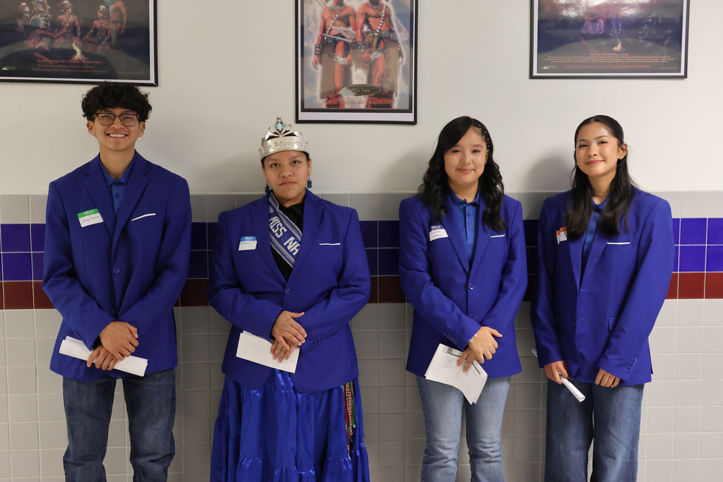 Four individuals in blue blazers stand together. One person wears a crown. Three hold papers.