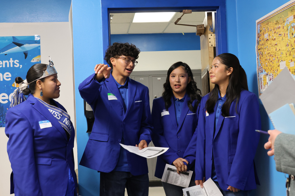 Four students in blue blazers and name tags discuss something in a hallway. A poster is on the wall.