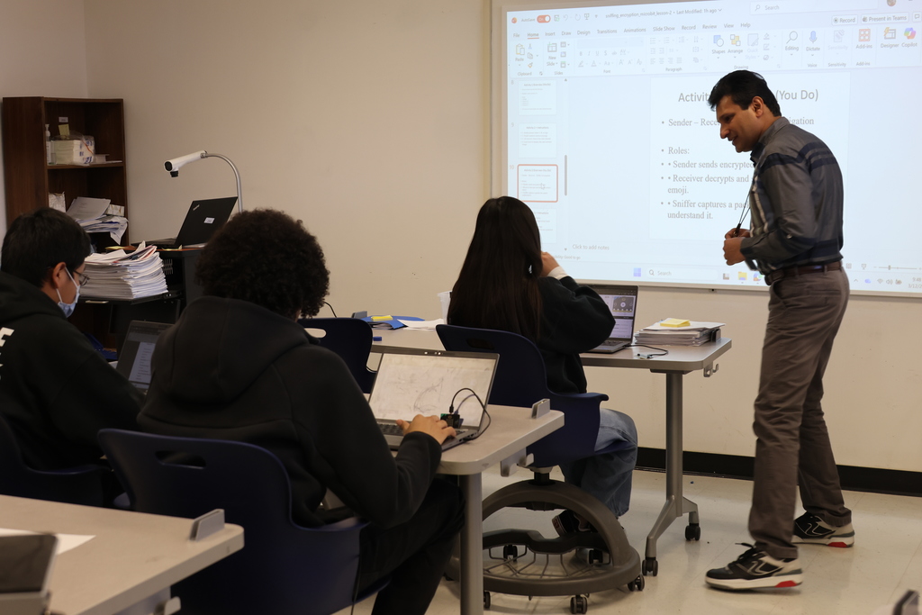 A classroom with a teacher standing at a desk. Students sit at tables, engaged with laptops.